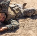 A military service member in MARPAT (Marine pattern) digital camouflage uniform and tactical vest is lying on sandy ground. The individual appears to be in a training or simulation scenario, possibly acting as a casualty role-player. The setting appears to be an outdoor training environment with sandy, dry terrain.