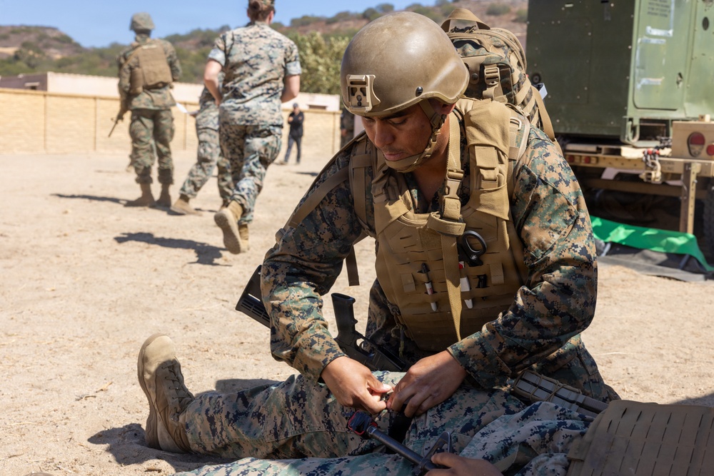 A U.S. Marine in full combat gear, including a helmet and tactical vest with backpack, is kneeling on sandy ground applying what appears to be a tourniquet or medical aid to a downed fellow Marine. The scene takes place at an outdoor training area, likely a military base, with other Marines walking in the background. The setting and actions suggest a combat casualty care or tactical combat casualty care (TCCC) training exercise.