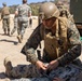 A U.S. Marine in full combat gear, including a helmet and tactical vest with backpack, is kneeling on sandy ground applying what appears to be a tourniquet or medical aid to a downed fellow Marine. The scene takes place at an outdoor training area, likely a military base, with other Marines walking in the background. The setting and actions suggest a combat casualty care or tactical combat casualty care (TCCC) training exercise.