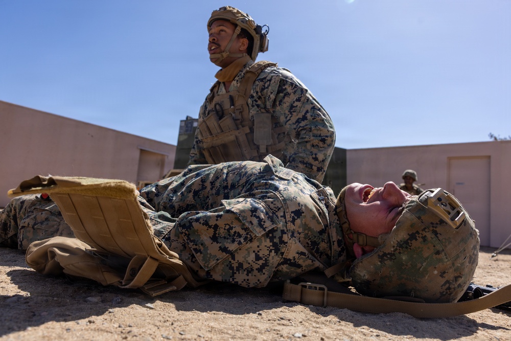 Two military personnel in MARPAT (Marine pattern) camouflage uniforms are engaged in what appears to be a combat casualty care or medical evacuation training exercise. One service member is lying on a litter/stretcher on sandy ground while another kneels behind them, appearing to provide care or prepare for transport. A third service member is visible in the background near a building structure.