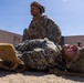 Two military personnel in MARPAT (Marine pattern) camouflage uniforms are engaged in what appears to be a combat casualty care or medical evacuation training exercise. One service member is lying on a litter/stretcher on sandy ground while another kneels behind them, appearing to provide care or prepare for transport. A third service member is visible in the background near a building structure.