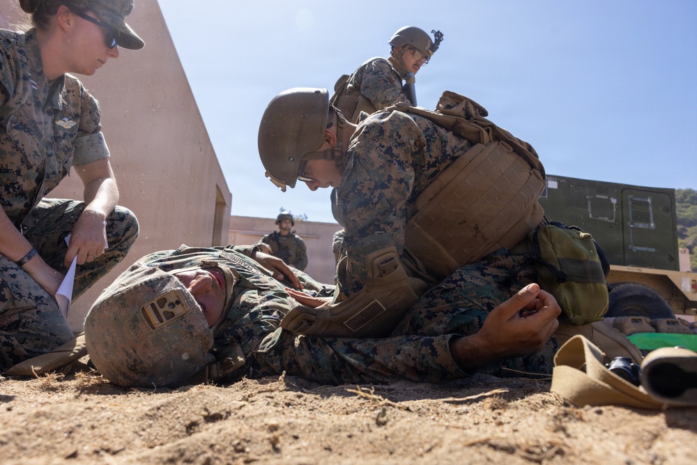 Military personnel in MARPAT (Marine pattern) camouflage uniforms are conducting what appears to be a combat casualty care or tactical combat casualty care (TCCC) training exercise. One service member is lying on the ground simulating a casualty while others attend to them in a field training environment. Multiple personnel are involved, suggesting a structured medical training drill.