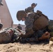 Military personnel in MARPAT (Marine pattern) camouflage uniforms are conducting what appears to be a combat casualty care or tactical combat casualty care (TCCC) training exercise. One service member is lying on the ground simulating a casualty while others attend to them in a field training environment. Multiple personnel are involved, suggesting a structured medical training drill.
