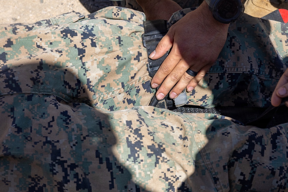 A close-up image showing a hand applying or adjusting a tourniquet on a person wearing US Marine Corps MARPAT digital camouflage uniform. The action appears to be a tactical combat casualty care (TCCC) or first aid procedure being performed on the lower torso/hip area. A black wristwatch is visible on the wrist of the person performing the procedure.