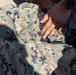 A close-up image showing a hand applying or adjusting a tourniquet on a person wearing US Marine Corps MARPAT digital camouflage uniform. The action appears to be a tactical combat casualty care (TCCC) or first aid procedure being performed on the lower torso/hip area. A black wristwatch is visible on the wrist of the person performing the procedure.