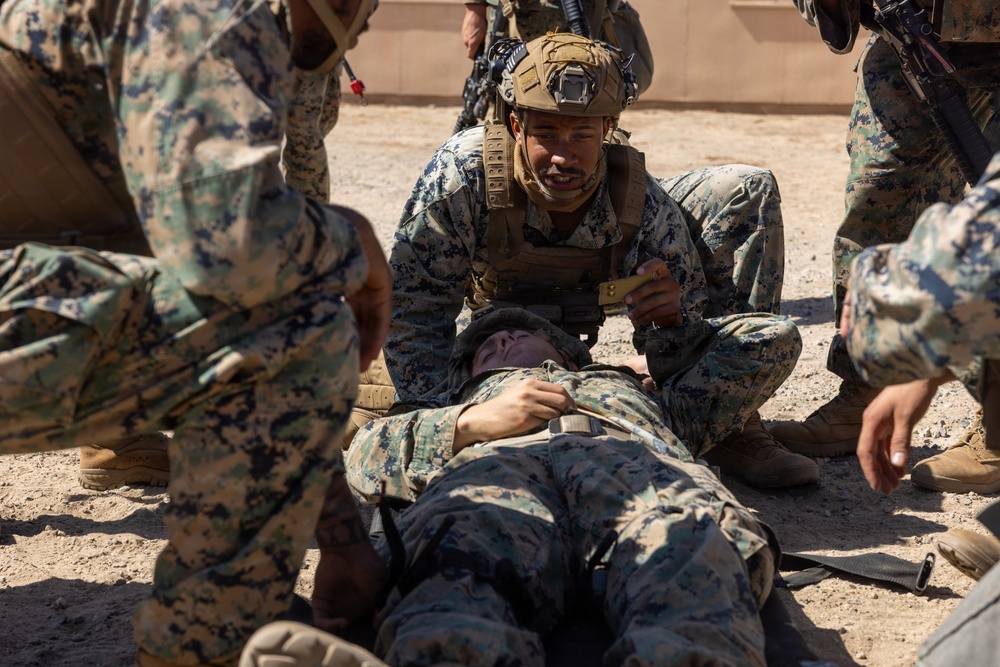 Military personnel in MARPAT (Marine pattern) camouflage uniforms are conducting what appears to be a combat casualty care or tactical combat casualty care (TCCC) training exercise. One service member wearing a tactical helmet with a camera mount is kneeling over a simulated casualty lying on the ground, while others assist. The setting appears to be an arid, outdoor training environment.