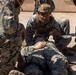Military personnel in MARPAT (Marine pattern) camouflage uniforms are conducting what appears to be a combat casualty care or tactical combat casualty care (TCCC) training exercise. One service member wearing a tactical helmet with a camera mount is kneeling over a simulated casualty lying on the ground, while others assist. The setting appears to be an arid, outdoor training environment.