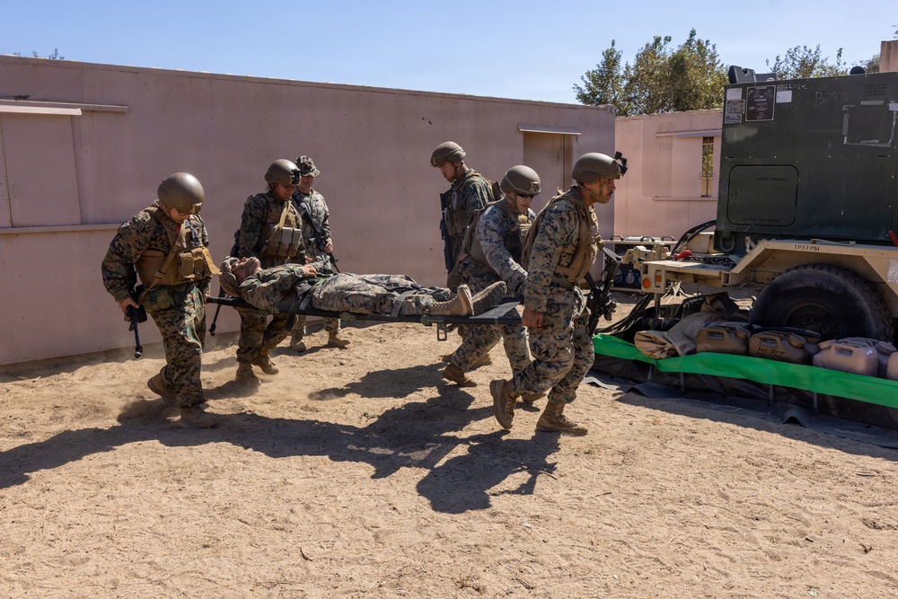 Military personnel in camouflage uniforms and helmets are carrying a person on a litter/stretcher toward a military vehicle. The scene appears to be a casualty evacuation drill or training exercise conducted in a desert-like environment near a tan/pink building. Multiple service members are coordinating the movement of the litter bearer toward what appears to be a military trailer or equipment platform.