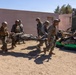 Military personnel in camouflage uniforms and helmets are carrying a person on a litter/stretcher toward a military vehicle. The scene appears to be a casualty evacuation drill or training exercise conducted in a desert-like environment near a tan/pink building. Multiple service members are coordinating the movement of the litter bearer toward what appears to be a military trailer or equipment platform.