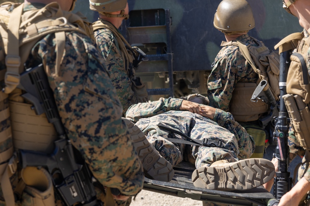 The image shows multiple military personnel in MARPAT (Marine pattern) camouflage uniforms attending to a fellow service member lying on a stretcher or litter. The scene appears to depict a casualty evacuation (CASEVAC) or medical training exercise, with soldiers loading or stabilizing the individual near a military vehicle. All personnel are equipped with tactical gear, body armor, and weapons.