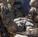 The image shows multiple military personnel in MARPAT (Marine pattern) camouflage uniforms attending to a fellow service member lying on a stretcher or litter. The scene appears to depict a casualty evacuation (CASEVAC) or medical training exercise, with soldiers loading or stabilizing the individual near a military vehicle. All personnel are equipped with tactical gear, body armor, and weapons.