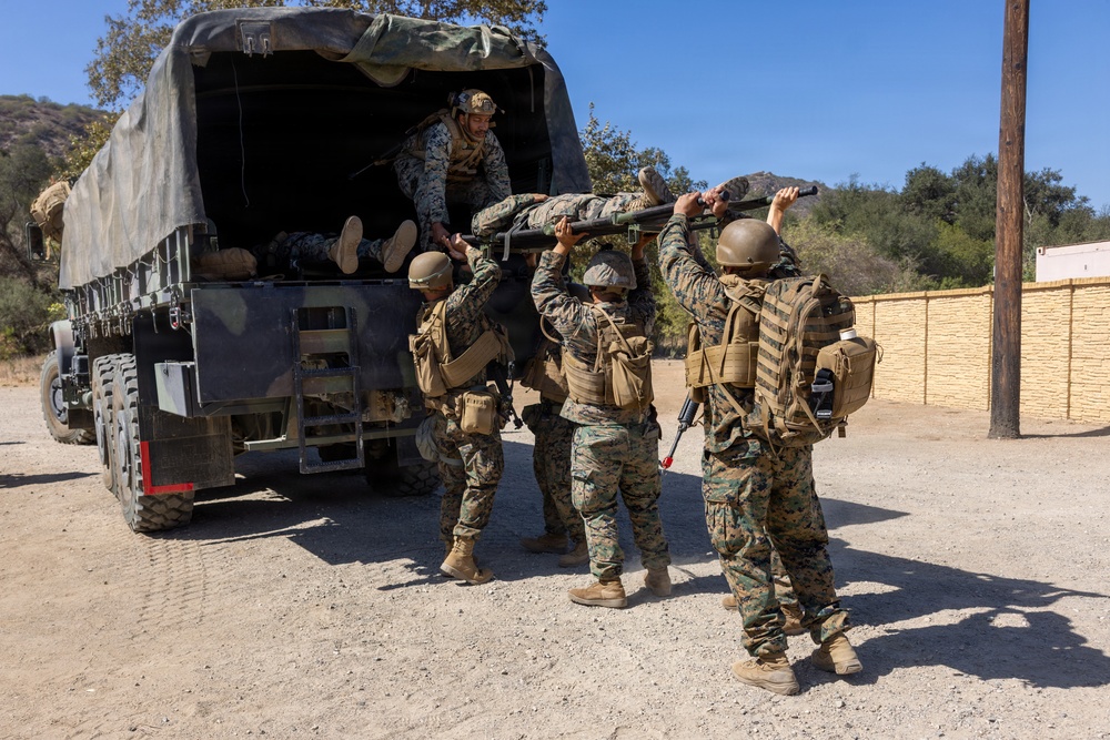 A group of military personnel in MARPAT camouflage uniforms are loading or unloading a casualty on a stretcher into/from a canvas-covered military truck. The scene appears to be a training exercise or field operation conducted in a dry, hilly terrain environment. At least four service members are actively involved in the casualty transport procedure.