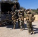 A group of military personnel in MARPAT camouflage uniforms are loading or unloading a casualty on a stretcher into/from a canvas-covered military truck. The scene appears to be a training exercise or field operation conducted in a dry, hilly terrain environment. At least four service members are actively involved in the casualty transport procedure.