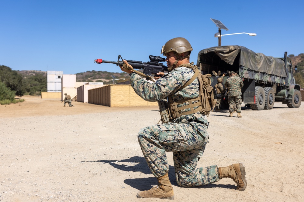 A military service member in MARPAT digital camouflage uniform is kneeling in a firing position, aiming a rifle equipped with a red training muzzle cap and optical scope. The setting appears to be a military training exercise at a base or training facility, with additional personnel and a large military cargo truck visible in the background. The environment is arid and dusty, consistent with a southwestern U.S. training installation.