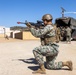 A military service member in MARPAT digital camouflage uniform is kneeling in a firing position, aiming a rifle equipped with a red training muzzle cap and optical scope. The setting appears to be a military training exercise at a base or training facility, with additional personnel and a large military cargo truck visible in the background. The environment is arid and dusty, consistent with a southwestern U.S. training installation.