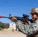 A military service member in digital camouflage (MARPAT pattern) is aiming an M16-style rifle equipped with a red training/MILES device on the muzzle and an optical scope. The setting appears to be a military training range with sandy terrain, a structure in the background, and another service member visible in the distance. The image captures a live training exercise in progress.