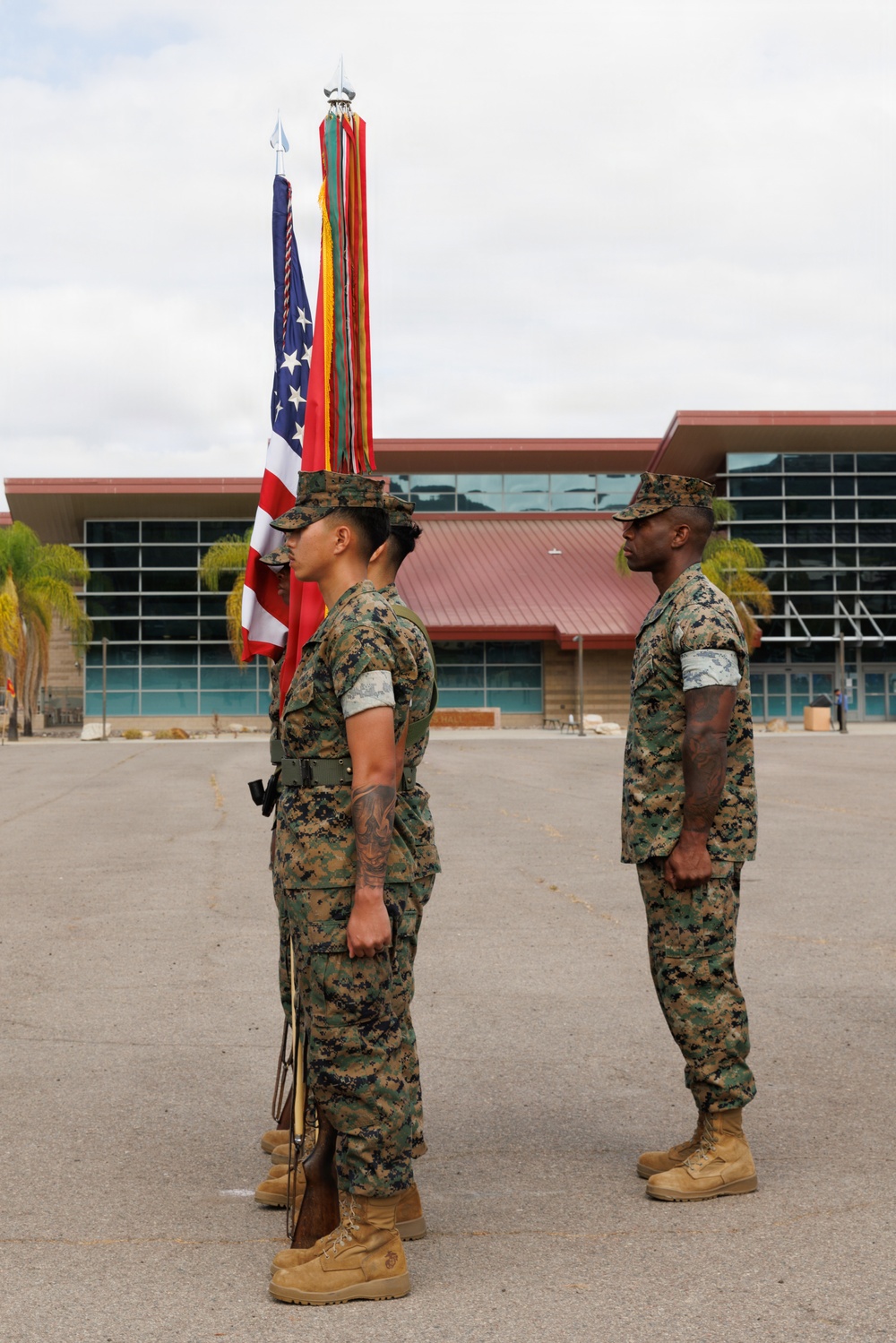 Three U.S. Marines in MARPAT woodland camouflage uniforms stand at attention during what appears to be a formal military ceremony or color guard formation. Two Marines in the foreground carry the U.S. national colors and what appears to be a unit guidon with streamers, while a third Marine stands at parade rest to the right. The setting is an outdoor parade deck in front of a large modern building, consistent with a military installation.