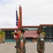 Three U.S. Marines in MARPAT woodland camouflage uniforms stand at attention during what appears to be a formal military ceremony or color guard formation. Two Marines in the foreground carry the U.S. national colors and what appears to be a unit guidon with streamers, while a third Marine stands at parade rest to the right. The setting is an outdoor parade deck in front of a large modern building, consistent with a military installation.