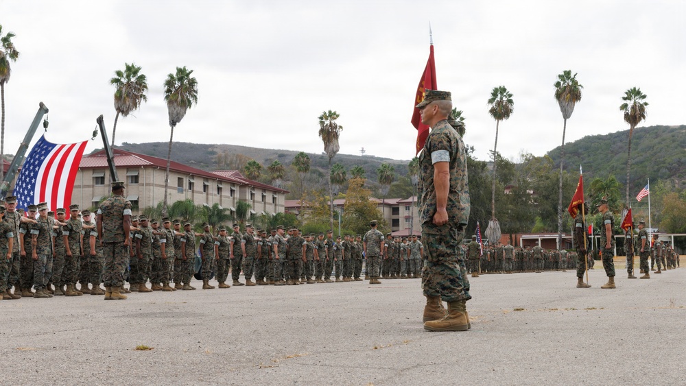 A formal U.S. Marine Corps military ceremony is taking place on a parade deck, likely a change of command or unit formation. Large formations of Marines in woodland MARPAT camouflage uniforms are standing at attention in organized ranks. The setting appears to be a Southern California military installation, identifiable by the palm trees, hillside terrain, and Spanish-style barracks buildings.