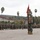 A formal U.S. Marine Corps military ceremony is taking place on a parade deck, likely a change of command or unit formation. Large formations of Marines in woodland MARPAT camouflage uniforms are standing at attention in organized ranks. The setting appears to be a Southern California military installation, identifiable by the palm trees, hillside terrain, and Spanish-style barracks buildings.