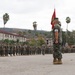 A formal U.S. Marine Corps military ceremony is taking place on a parade deck, likely a change of command or unit formation. Large formations of Marines in woodland MARPAT camouflage uniforms are standing at attention in organized ranks. The setting appears to be a Southern California military installation, identifiable by the palm trees, hillside terrain, and Spanish-style barracks buildings.