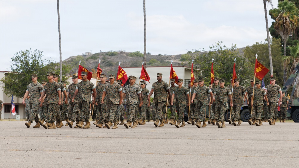 A formation of U.S. Marines in woodland MARPAT camouflage uniforms marches across a parade deck during what appears to be a formal military ceremony or review. Multiple guidon bearers carry red FMF (Fleet Marine Force) pennants on staffs with spear-point finials. The setting appears to be a Southern California military installation, consistent with Camp Pendleton or a similar base, based on the terrain and vegetation.