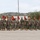 A formation of U.S. Marines in woodland MARPAT camouflage uniforms marches across a parade deck during what appears to be a formal military ceremony or review. Multiple guidon bearers carry red FMF (Fleet Marine Force) pennants on staffs with spear-point finials. The setting appears to be a Southern California military installation, consistent with Camp Pendleton or a similar base, based on the terrain and vegetation.