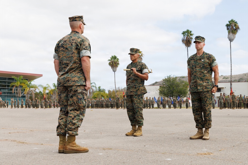 A formal U.S. Marine Corps military ceremony is taking place on a parade deck, likely a change of command or similar official event. Three Marines in the foreground are in prominent positions, with one holding what appears to be a ceremonial sword or saber. A large formation of Marines stands at attention in the background.