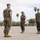 A formal U.S. Marine Corps military ceremony is taking place on a parade deck, likely a change of command or similar official event. Three Marines in the foreground are in prominent positions, with one holding what appears to be a ceremonial sword or saber. A large formation of Marines stands at attention in the background.