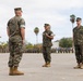 A formal U.S. Marine Corps military ceremony is taking place on a parade deck, likely a change of command or similar official event. Three Marines in the foreground are in prominent positions, with one holding what appears to be a ceremonial sword or saber. A large formation of Marines stands at attention in the background.
