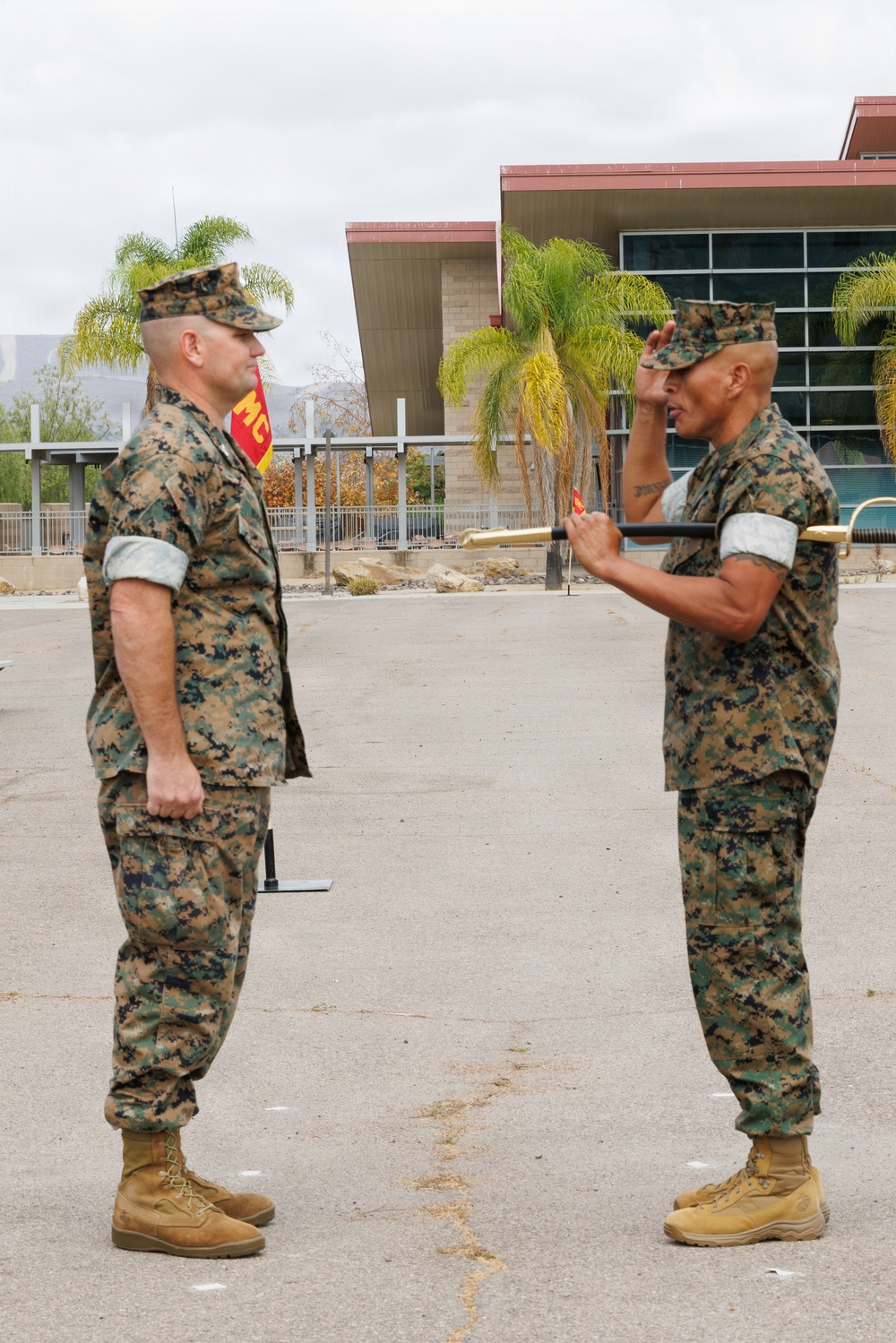 Two U.S. Marines in MARPAT woodland camouflage uniforms are engaged in what appears to be a formal military ceremony, likely a change of command or award presentation. The Marine on the right is rendering a salute while holding what appears to be a ceremonial guidon or award case, facing the Marine on the left who stands at attention. The setting appears to be a military installation, consistent with a base in a warm climate given the palm trees and building architecture.