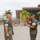Two U.S. Marines in MARPAT woodland camouflage uniforms are engaged in what appears to be a formal military ceremony, likely a change of command or award presentation. The Marine on the right is rendering a salute while holding what appears to be a ceremonial guidon or award case, facing the Marine on the left who stands at attention. The setting appears to be a military installation, consistent with a base in a warm climate given the palm trees and building architecture.