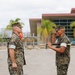 Two U.S. Marines in MARPAT woodland camouflage uniforms are engaged in what appears to be a formal military ceremony, likely a change of command or award presentation. The Marine on the right is rendering a salute while holding what appears to be a ceremonial guidon or award case, facing the Marine on the left who stands at attention. The setting appears to be a military installation, consistent with a base in a warm climate given the palm trees and building architecture.