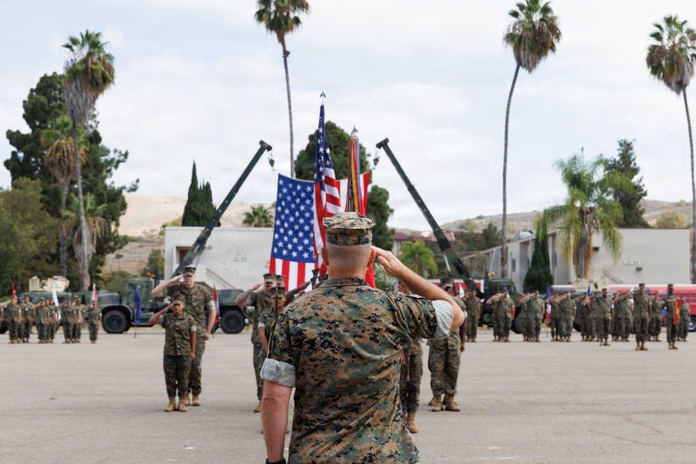 A U.S. Marine Corps military ceremony is taking place on a parade deck, with a senior Marine in the foreground rendering a salute toward a color guard bearing the American flag and additional unit colors. Formations of Marines in MARPAT digital camouflage uniforms are standing at attention or saluting in organized ranks. The setting appears to be a military installation consistent with a Southern California base, likely Camp Pendleton or a similar facility, indicated by the palm trees, arid hills, and base infrastructure.