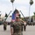 A U.S. Marine Corps military ceremony is taking place on a parade deck, with a senior Marine in the foreground rendering a salute toward a color guard bearing the American flag and additional unit colors. Formations of Marines in MARPAT digital camouflage uniforms are standing at attention or saluting in organized ranks. The setting appears to be a military installation consistent with a Southern California base, likely Camp Pendleton or a similar facility, indicated by the palm trees, arid hills, and base infrastructure.