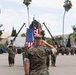 A U.S. Marine Corps military ceremony is taking place on a parade deck, with a senior Marine in the foreground rendering a salute toward a color guard bearing the American flag and additional unit colors. Formations of Marines in MARPAT digital camouflage uniforms are standing at attention or saluting in organized ranks. The setting appears to be a military installation consistent with a Southern California base, likely Camp Pendleton or a similar facility, indicated by the palm trees, arid hills, and base infrastructure.