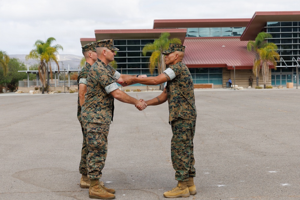 Two military personnel in U.S. Marine Corps MARPAT woodland camouflage uniforms are shaking hands on a paved parade deck or parking area. The setting appears to be a military installation, likely in a warm, arid region based on the palm trees and mountain backdrop. The handshake suggests a formal military ceremony, possibly a change of command, promotion, or award presentation.