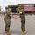Two military personnel in U.S. Marine Corps MARPAT woodland camouflage uniforms are shaking hands on a paved parade deck or parking area. The setting appears to be a military installation, likely in a warm, arid region based on the palm trees and mountain backdrop. The handshake suggests a formal military ceremony, possibly a change of command, promotion, or award presentation.