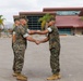 Two military personnel in U.S. Marine Corps MARPAT woodland camouflage uniforms are shaking hands on a paved parade deck or parking area. The setting appears to be a military installation, likely in a warm, arid region based on the palm trees and mountain backdrop. The handshake suggests a formal military ceremony, possibly a change of command, promotion, or award presentation.