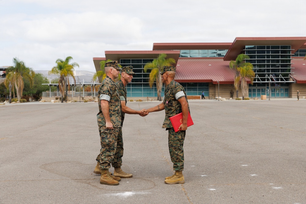 Three military personnel in U.S. Marine Corps MARPAT camouflage uniforms are gathered on a paved parade deck or parking area. Two of the individuals are shaking hands in what appears to be a formal ceremony or recognition event, with one holding a red folder or award binder. The setting appears to be a military installation, likely on the West Coast based on the architecture and palm trees.