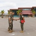 Three military personnel in U.S. Marine Corps MARPAT camouflage uniforms are gathered on a paved parade deck or parking area. Two of the individuals are shaking hands in what appears to be a formal ceremony or recognition event, with one holding a red folder or award binder. The setting appears to be a military installation, likely on the West Coast based on the architecture and palm trees.
