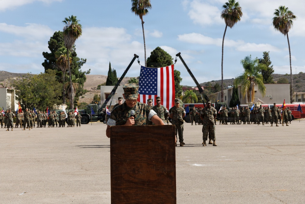 A U.S. Marine in camouflage uniform and cover speaks at a wooden podium during a formal military ceremony on a parade deck. An American flag is displayed prominently in the background, suspended between two angled poles, with formations of Marines standing at attention on both sides. The setting appears to be a military installation, likely on the West Coast based on the palm trees and terrain.