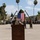 A U.S. Marine in camouflage uniform and cover speaks at a wooden podium during a formal military ceremony on a parade deck. An American flag is displayed prominently in the background, suspended between two angled poles, with formations of Marines standing at attention on both sides. The setting appears to be a military installation, likely on the West Coast based on the palm trees and terrain.