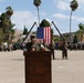 A U.S. Marine in camouflage uniform and cover speaks at a wooden podium during a formal military ceremony on a parade deck. An American flag is displayed prominently in the background, suspended between two angled poles, with formations of Marines standing at attention on both sides. The setting appears to be a military installation, likely on the West Coast based on the palm trees and terrain.
