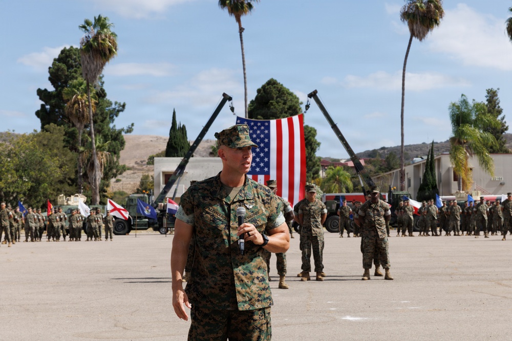 A U.S. Marine Corps officer in MARPAT camouflage uniform is speaking into a microphone at what appears to be a formal military ceremony or parade. A large American flag is suspended between two cranes in the background, with formations of Marines standing at attention on a parade deck. The setting appears to be a military installation, likely in Southern California based on the terrain and palm trees.