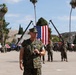 A U.S. Marine Corps officer in MARPAT camouflage uniform is speaking into a microphone at what appears to be a formal military ceremony or parade. A large American flag is suspended between two cranes in the background, with formations of Marines standing at attention on a parade deck. The setting appears to be a military installation, likely in Southern California based on the terrain and palm trees.