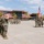A U.S. Marine Corps military ceremony is taking place on a parade deck, likely at a Southern California base given the architecture, palm trees, and terrain. Marines in MARPAT camouflage uniforms are marching in formation while officers render salutes. A color guard carries the U.S. national flag and a red Marine Corps unit guidon.