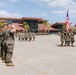 A U.S. Marine Corps military ceremony is taking place on a parade deck, likely at a Southern California base given the architecture, palm trees, and terrain. Marines in MARPAT camouflage uniforms are marching in formation while officers render salutes. A color guard carries the U.S. national flag and a red Marine Corps unit guidon.