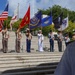 A joint military color guard ceremony is taking place on the steps of what appears to be a memorial or government building. Personnel from multiple U.S. military branches are standing in formation, each holding their respective service flags. A senior officer in the foreground, viewed from behind, appears to be presiding over or observing the ceremony.