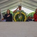 A formal military memorial ceremony is taking place under a large white tent, likely at a military cemetery or memorial site. Four individuals stand at attention behind a stone monument or altar, with a floral wreath of yellow and white flowers prominently displayed. Three civilians and one uniformed military officer are participating, with attendees in military dress uniforms seated in the background.
